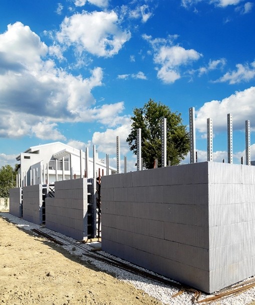 Cantiere con struttura in costruzione utilizzando il sistema costruttivo Bioisotherm. Blocchi di casseforme in polistirene espanso sono assemblati per creare pareti di edifici antisismici, offrendo isolamento termico e acustico. Sfondo di cielo azzurro con nuvole.