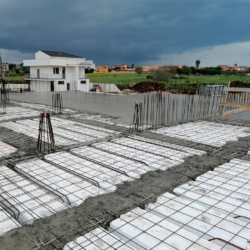 Costruzione di un edificio in calcestruzzo armato con sistema Bioisotherm. Sono visibili casseforme in polistirene espanso e armature in acciaio su una superficie in costruzione. Sullo sfondo, un edificio completato e un cielo nuvoloso.