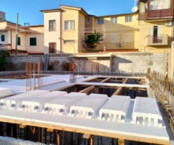 Costruzione di un edificio residenziale in fase iniziale con solai in polistirolo Bioisotherm. Sullo sfondo, case gialle e arancioni. Il sistema di isolamento termico Bioisotherm è visibile nei pannelli bianchi, migliorando l'efficienza energetica della struttura.
