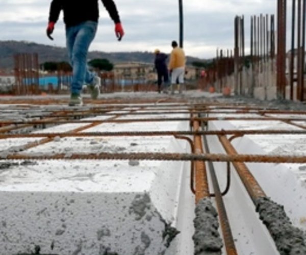 Operai camminano su un cantiere edile dove si utilizza il sistema costruttivo Bioisotherm. Struttura in calcestruzzo con barre d’acciaio e pannelli isolanti in polistirolo. Sfondo di colline e cielo nuvoloso, evidenziando l'efficienza e l'innovazione nel settore delle costruzioni.