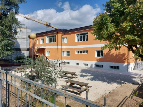 Edificio scolastico a due piani con facciata arancione e bianca, recentemente ristrutturato con tecnologia a cappotto sismico Argisol. Presenti tavoli da picnic nel cortile, alberi e una gru sullo sfondo. Progetto di adeguamento sismico ed energetico a Ascoli Piceno, con Bioisotherm.
